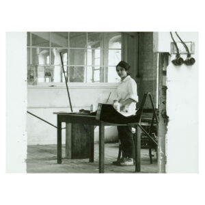 Black and white photo of Eva Hesse standing in front of a work table, looking to the camera, in her studio in Kettwig an der Ruhr, Germany.