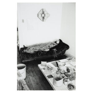Black and white photo of Eva Hesse lying on a sofa in her studio in Bowery, New York in 1969, covered by ropes. Work table with materials in the foreground.
