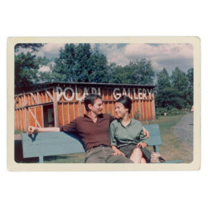Colored photograph showing Eva Hesse (on the right) and Tom Doyle (on the left) sitting on a bench a looking at each other in Wookstock in 1962.