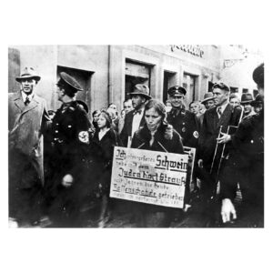 Black and white photo of S.S. men leading a woman through the streets wearing a sign that reads: “I am a swine. I slept with a Jew, Karl Strauss, and thus polluted the German race.” The photo was taken in Kulmbach, Germany, in 1938.